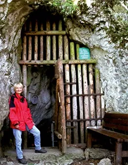 Eine Frau in einer roten Jacke steht vor einem Holztor in einer Höhle, mit einer Bank rechts von ihr und einem grünen Schild über dem Tor.