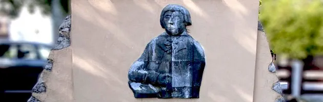 Eine verwitterte Statue eines Mannes in Uniform ist an einer beigen Wand befestigt. Die Statue ist blau und scheint aus Stein oder Metall zu sein.