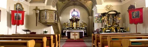 Ein Innenraum einer Kirche mit hölzernen Kirchenbänken, einem zentralen Altar und dekorativen Elementen, einschließlich Bannern und religiösen Symbolen.
