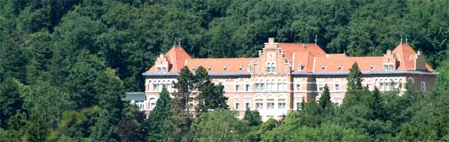 Ein großes Gebäude mit roten Dächern und vielen Fenstern ist von dichten grünen Bäumen umgeben.