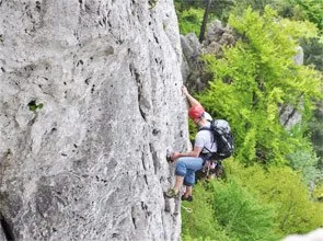 Eine Person klettert mit rotem Helm, Rucksack und Gurtzeug. Sie greift eine Felswand, mit einem Wald im Hintergrund.