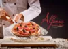 Ein Koch in einer weißen Uniform schneidet eine Pizza mit schwarzen Oliven, roten Tomaten und anderen Toppings auf einem Holzbrett.