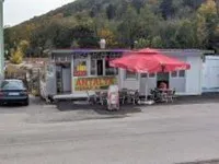 Ein kleiner Imbisswagen mit einem roten Schirm steht auf einem Parkplatz mit einem Berg im Hintergrund.