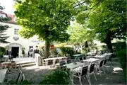 Ein Außenbereich zum Essen mit Tischen und Stühlen, umgeben von üppigen grünen Bäumen und einem weißen Gebäude im Hintergrund.