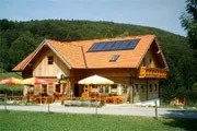Ein Chalet-Haus mit Solarpanelen auf dem Dach, umgeben von einem üppigen grünen Feld und Bergen im Hintergrund. Vor dem Haus stehen Außenschirme und Tische.