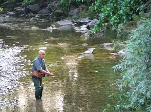 Ein älterer Mann steht in einem flachen Fluss und zeigt auf etwas im Wasser. Er trägt ein blaues Shirt, grüne Hosen und Stiefel. Felsen und Grünpflanzen säumen die Flussufer.