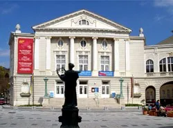 Ein klassisches Gebäude mit einer Statue davor, großen Fenstern und Bannern an der Seite. Der Himmel ist klar und blau.