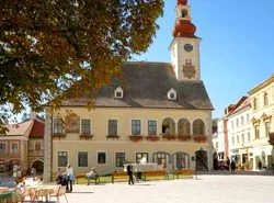 Ein großes historisches Gebäude mit einem Glockenturm steht in der Mitte eines Stadtplatzes, umgeben von Bänken, Tischen und Menschen, die spazieren gehen.