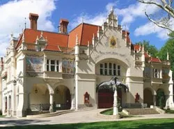 Ein großes, kunstvoll gestaltetes Gebäude mit rotem Dach und weißen Wänden, mit mehreren Bogenfenstern und Türen, in einer üppigen grünen Landschaft mit einem Brunnen im Vordergrund.