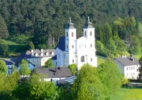 Luftaufnahme einer großen weißen Kirche mit zwei Türmen, umgeben von üppigem grünem Baumbestand und Gebäuden in einem bergigen Gebiet.