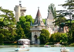 Ein ruhiger See mit mehreren kleinen Booten, umgeben von üppigem Grün und einer schlossartigen Struktur im Hintergrund.
