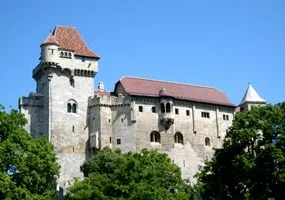 Eine alte Burg mit rotem Dach und mehreren Türmen, umgeben von üppigem grünem Laub unter einem klaren blauen Himmel.