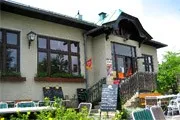 Ein weißes Gebäude mit schwarzen Akzenten und großen Fenstern, das einen Garten mit Pflanzen und einer Treppe zum Eingang aufweist. Draußen stehen Tische und Stühle sowie eine Tafel mit Schrift.