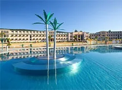 Ein großer Swimmingpool mit einer zentralen Palmenbaumdekoration, umgeben von einem Resort mit mehreren Gebäuden unter einem klaren blauen Himmel.
