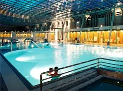 Ein überdachter Innenpool mit Glasdach, der einen großen Poolbereich, einen kleineren Pool und zwei schwimmende Personen zeigt. Der Pool ist gut beleuchtet, und es gibt Liegestühle und eine Brücke im Hintergrund.