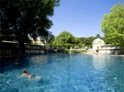 Eine Person schwimmt in einem großen Außenpool, umgeben von üppiger Grünfläche und klarem blauem Himmel.