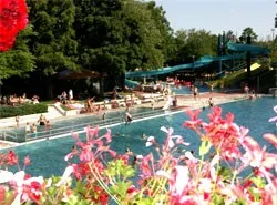Ein großer Außenpool mit schwimmenden und entspannenden Menschen, umgeben von Bäumen, Blumen und einer Wasserrutsche.