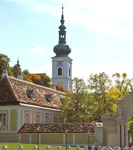 Ein historisches Gebäude mit einem hohen Turm und einer Uhr, umgeben von Bäumen und einem Zaun, unter einem klaren blauen Himmel.
