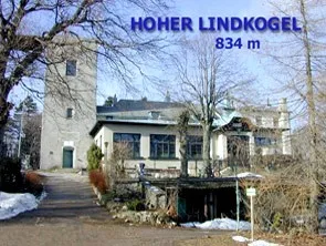 Ein großes weißes Gebäude namens 'Hoher Lindkogel' mit einem Turm und umgeben von Bäumen, auf einer Höhe von 834 Metern gelegen.