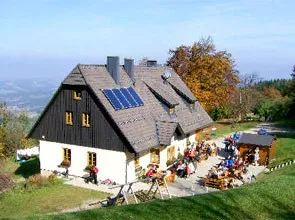 Ein Haus mit Solarpanelen auf dem Dach, umgeben von einer Gruppe von Menschen, die draußen an Tischen sitzen, in einem Grasbereich mit Bäumen im Hintergrund.