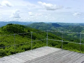 Eine Holzterrasse mit einem Metallgeländer bietet einen Panoramablick auf üppige grüne Berge unter einem teilweise bewölkten Himmel.