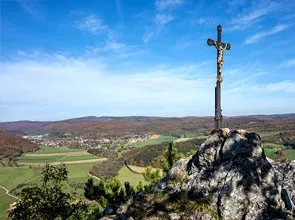 Ein Kreuz steht hoch auf einem felsigen Hügel und bietet einen Panoramablick auf ein üppiges Tal darunter unter einem klaren blauen Himmel.