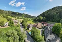 Eine Luftaufnahme eines kleinen Dorfes, umgeben von üppigen grünen Hügeln und dichten Wäldern unter einem klaren blauen Himmel.