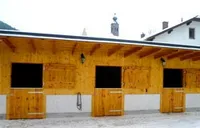 Ein Holzstall mit drei Ställen, jeder mit einer Holztür und einem Fenster, unter einem geneigten Dach mit einem Schornstein oben, umgeben von einer verschneiten Landschaft.