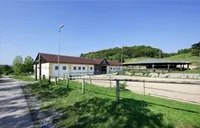 Ein großes weißes Gebäude mit einem braunen Dach steht neben einem eingezäunten Bereich mit einem Grasfeld und einer Straße. Bäume und ein Hügel sind im Hintergrund sichtbar.