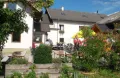 Ein weißes Haus mit grauem Dach und einem Garten davor, mit Menschen, die unter einem gelben Schirm draußen sitzen.