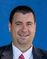 Ein Mann in Anzug und weißem Hemd lächelt für ein Nahaufnahmefoto vor blauem Hintergrund.