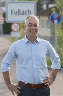 Ein Mann steht mit den Händen auf den Hüften, trägt ein hellblaues Hemd und einen braunen Gürtel. Er lächelt und posiert wahrscheinlich für ein Foto. Hinter ihm befindet sich ein Straßenschild mit dem Wort FuBach und weiteren Schildern.