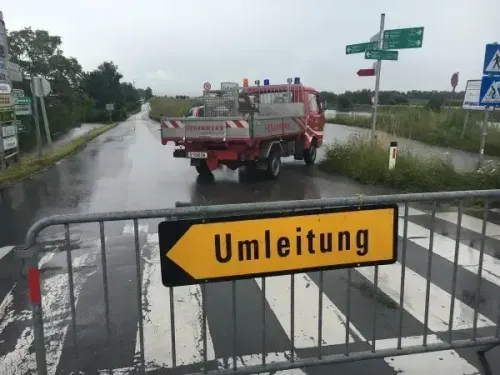 Ein roter LKW fährt auf einer nassen Straße mit einer Fußgängerampel. Ein gelbes Schild mit schwarzem Pfeil und der Aufschrift 'Umleitung' ist an der Schranke befestigt. Straßenschilder und Stangen befinden sich am Straßenrand. Das Gebiet ist von Gras und Bäumen umgeben.