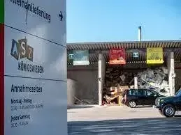Ein Schild für Abfallwirtschaftsbetrieb mit AS7-Logo, in der Nähe eines Depots mit einem Haufen Abfall. Zwei Autos sind vor dem Depot geparkt, mit einem klaren Himmel im Hintergrund.