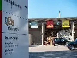 Ein Schild für Abfallwirtschaftsbetrieb mit AS7-Logo, in der Nähe eines Depots mit einem Haufen Abfall. Zwei Autos sind vor dem Depot geparkt, mit einem klaren Himmel im Hintergrund.