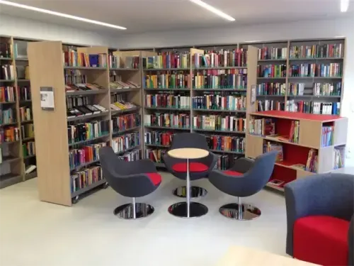 Eine leere Bibliothek mit weißem Boden und einigen Stühlen um einen Tisch in der Mitte. Es gibt Regale voller Bücher auf beiden Seiten des Raumes.