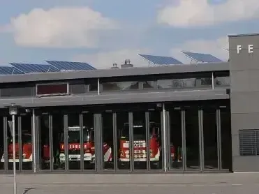 Eine moderne Feuerwache mit Solarpanelen auf dem Dach. Im Inneren sind drei rote Feuerwehrfahrzeuge in der Garage geparkt. Das Gebäude hat eine Glasfassade und ein Schild mit den Buchstaben 'FE'.