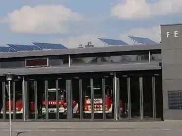 Eine moderne Feuerwache mit Solarpanelen auf dem Dach. Im Inneren sind drei rote Feuerwehrfahrzeuge in der Garage geparkt. Das Gebäude hat eine Glasfassade und ein Schild mit den Buchstaben 'FE'.