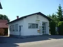 Ein weißes Gebäude mit schwarzem Dach und mehreren Fenstern steht auf einem Parkplatz. Bäume und Sträucher sind im Hintergrund sichtbar.