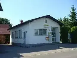 Ein weißes Gebäude mit schwarzem Dach und mehreren Fenstern steht auf einem Parkplatz. Bäume und Sträucher sind im Hintergrund sichtbar.