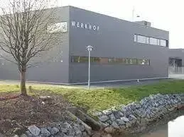Ein graues Gebäude namens Werkhof mit mehreren Fenstern und einem Laternenpfosten davor. Es ist von Gras, Steinen und einem kleinen Bach umgeben.