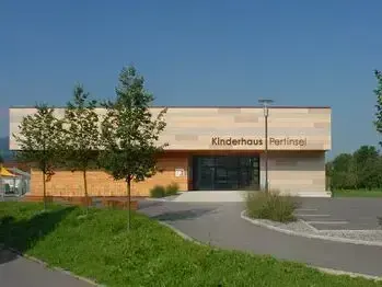 Ein modernes Gebäude mit der Aufschrift Kinderhaus Perlingei, gelegen in einem grasigen Bereich mit Bäumen und Pflanzen. Das Gebäude hat einen großen Eingang und einen Parkplatz davor.