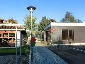 Eine Person steht auf einem gepflasterten Weg vor einem modernen Gebäude mit Glasfenstern, unter einer hohen Straßenlampe, mit Bäumen und einem Grasbereich im Hintergrund.