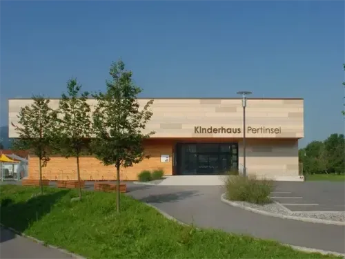 Das Kinderhaus Pertinsel-Gebäude hat eine moderne Fassade mit einem Rasen und Bäumen davor. Der Eingang hat große Glastüren und Fenster.
