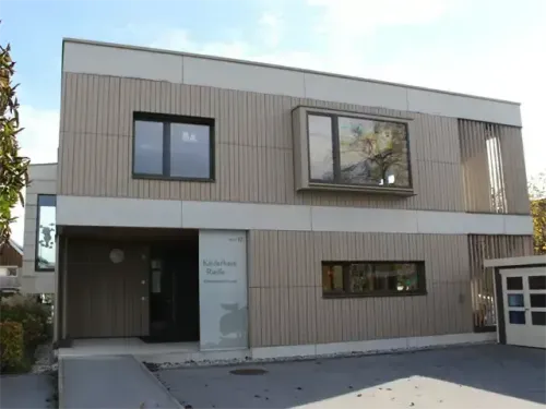 Das Bild zeigt ein modernes Gebäude mit grauer Außenfassade, großen Fenstern und einem zentralen Eingang. Das Schild lautet 'Kinderhaus Riedel'.