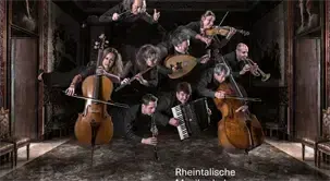 Eine Gruppe von Musikern spielt drinnen. Sie spielen verschiedene Instrumente wie Violinen, Celli und Flöten. Einige Musiker stehen, andere sitzen auf dem Boden. Der Raum hat Holzwände und einen Teppichboden. Der Text 'Rheintalische' steht unten rechts.