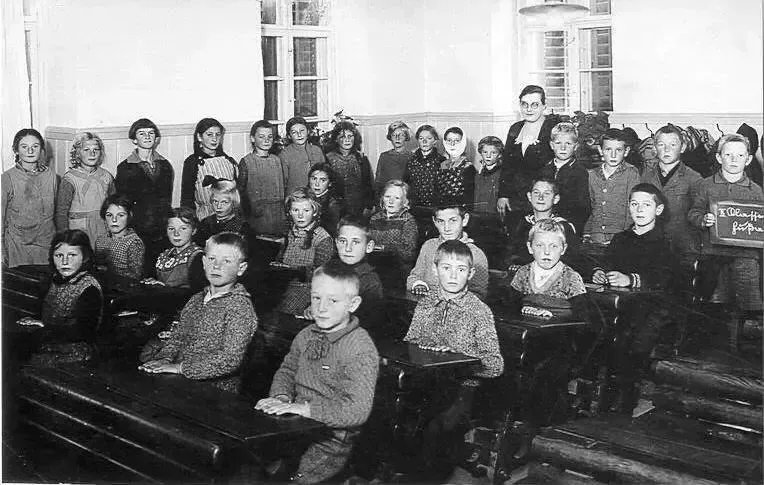Ein Schwarz-Weiß-Foto einer Klasse mit Schülern und einer Lehrerin, alle in Winterkleidung, schauen in die Kamera, einige mit erhobenen Händen.