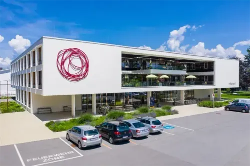 Ein modernes weißes Gebäude mit rotem Logo zeigt eine Außenansicht, mit einem Parkplatz, auf dem Autos geparkt sind, einer Terrasse mit Sonnenschirmen und viel Grün.