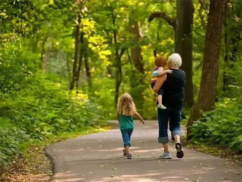 Eine ältere Frau geht auf einem Pfad in einem Park, trägt ein kleines Kind auf der Schulter, während ein anderes Kind neben ihr geht.