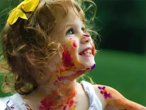 Ein kleines Mädchen mit einer gelben Schleife im Haar lächelt mit Farbe im Gesicht und an den Armen.